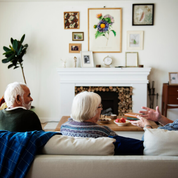 Cztery starsze osoby siedzą razem w jasnym salonie — trzy na kanapie i jedna w fotelu — rozmawiają przy stoliku kawowym, w tle kominek z dekoracjami i obrazy na ścianie.
