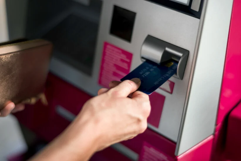 Osoba wkłada kartę bankową do bankomatu trzymając w drugiej ręce portfel.