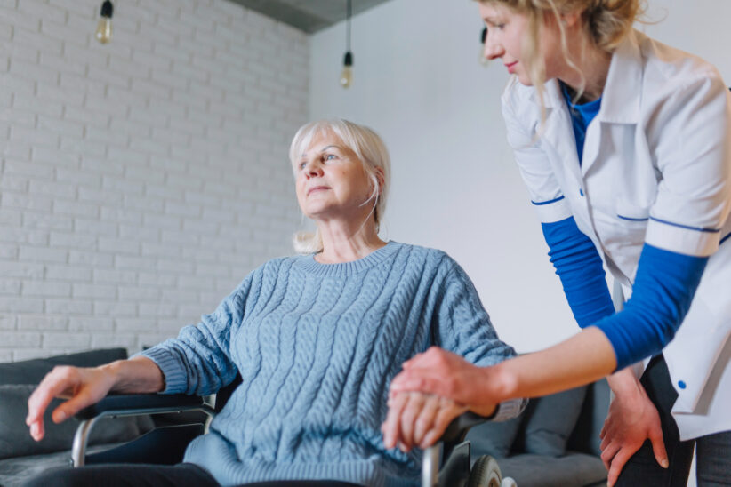seniorka siedzi na wózku, obok niej stoi pielęgniarka