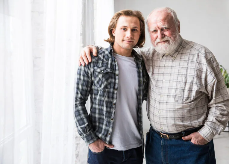 Młody mężczyzna stoi obok starszego mężczyzny, obejmują się ramionami i patrzą w obiektyw aparatu.