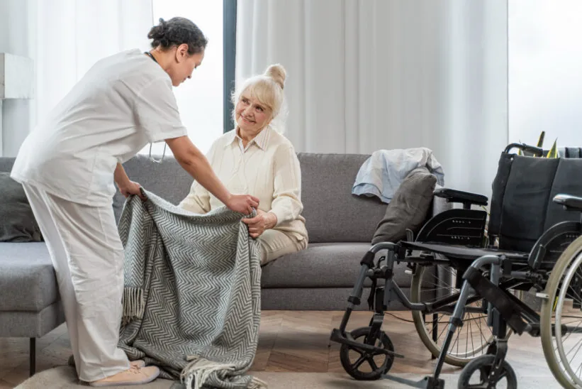 seniorka siedzi na łóżku, opiekunka jej pomaga
