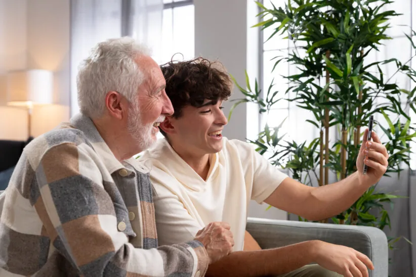 Starszy mężczyzna i nastolatek siedzą razem na kanapie i uśmiechnięci robią selfie smartfonem.