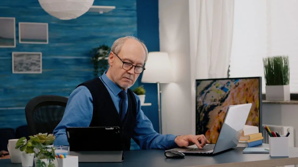 Starszy mężczyzna siedzi przy biurku w domowym gabinecie, pracuje na laptopie, a obok ma tablet i notatki.