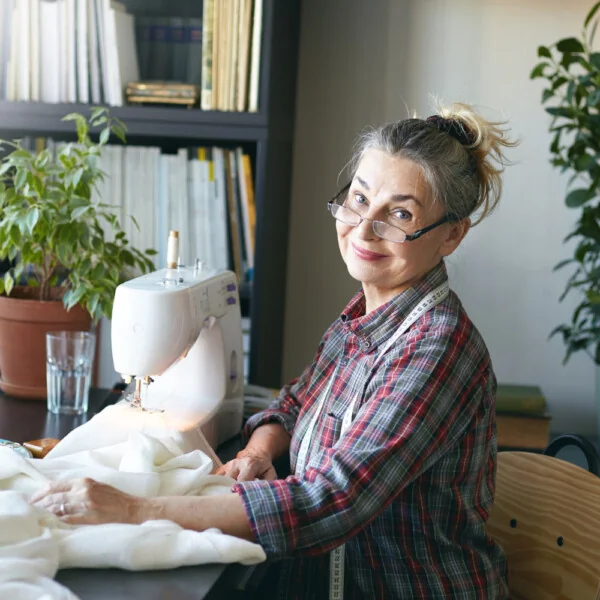 Starsza kobieta siedzi przy stole i szyje na maszynie, uśmiechając się do aparatu, w tle widać rośliny i regał z książkami.