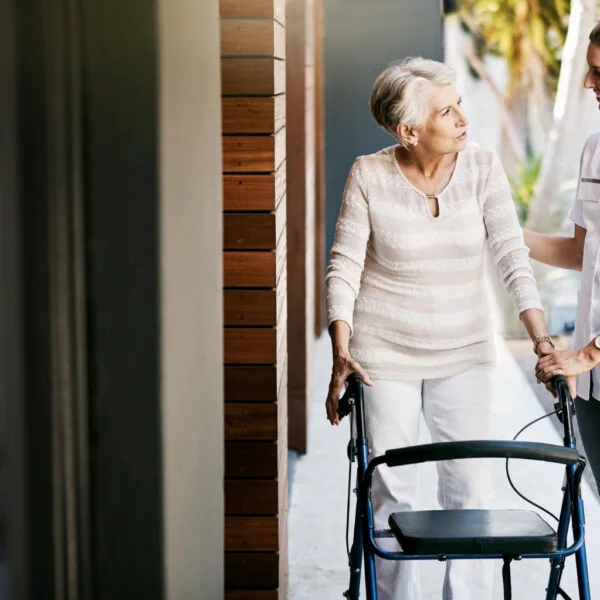 Starsza kobieta z balkonikiem spaceruje na zewnątrz w towarzystwie opiekunki, która wspiera ją i rozmawia z uśmiechem