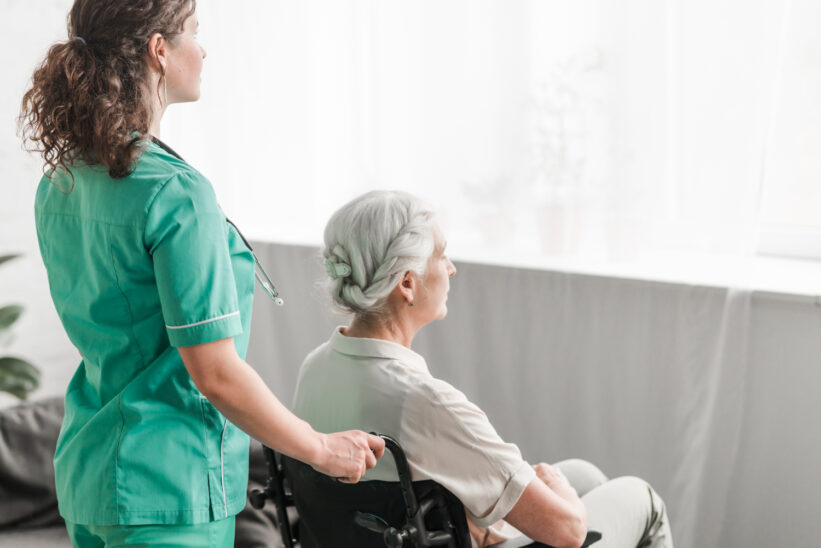 Opiekunka prowadzi seniorkę na wózku inwalidzkim w jasnym pokoju domu opieki.