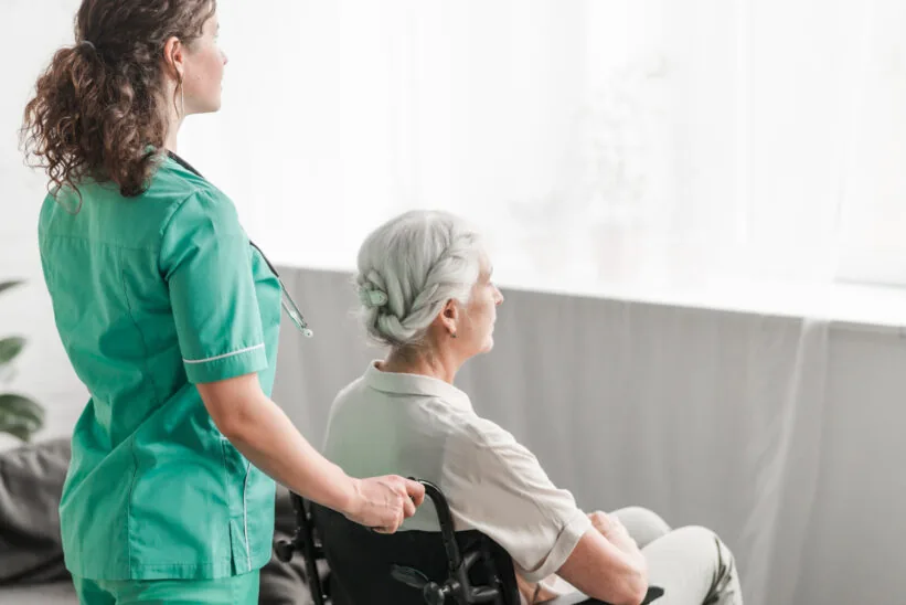Opiekunka prowadzi seniorkę na wózku inwalidzkim w jasnym pokoju domu opieki.