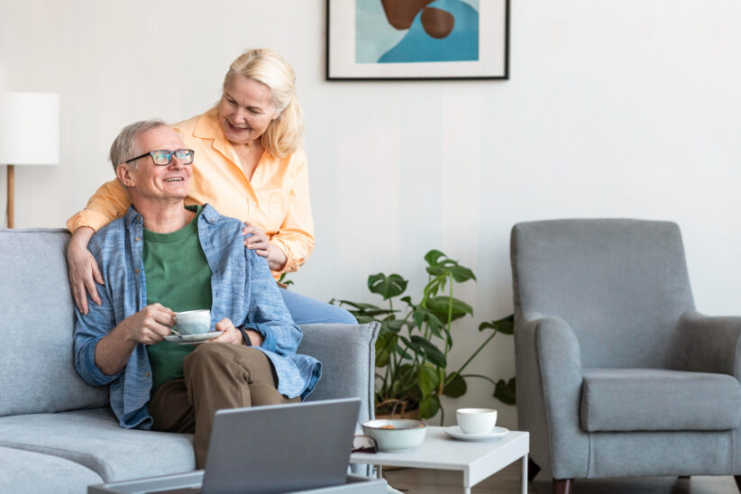 Uśmiechnięta para seniorów spędza czas w nowoczesnym salonie. Mężczyzna siedzi na kanapie z filiżanką kawy, a kobieta stoi obok niego