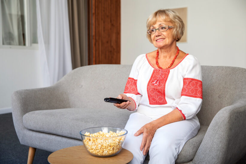 Seniorka siedzi na kanapie z pilotem w dłoni i ogląda telewizję w domowym salonie.