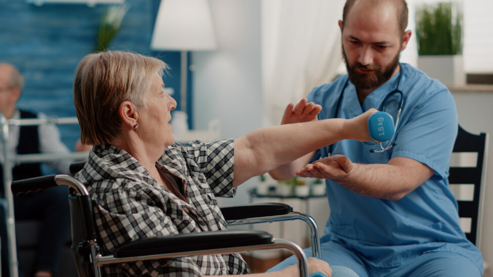 Seniorka wykonująca ćwiczenie z hantlem pod opieką rehabilitanta podczas terapii ruchowej.
