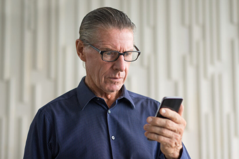 Senior w okularach patrzy z niepokojem na telefon komórkowy, podejrzewając oszustwo bankowe.