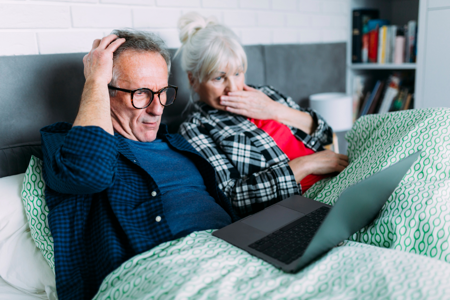 Zaniepokojona para seniorów leży w łóżku i patrzy na ekran laptopa.