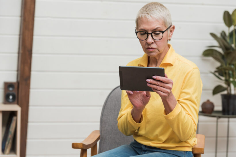 Kobieta seniorka w żółtej bluzce korzysta z tabletu w domu