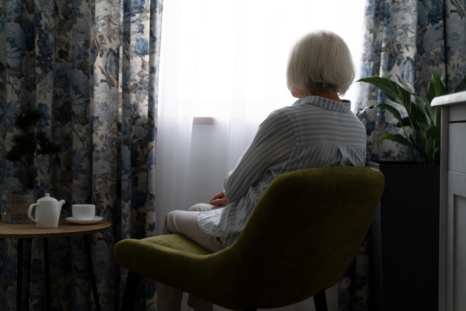 Seniorka siedząca samotnie przy oknie w pokoju domu opieki lub mieszkania.