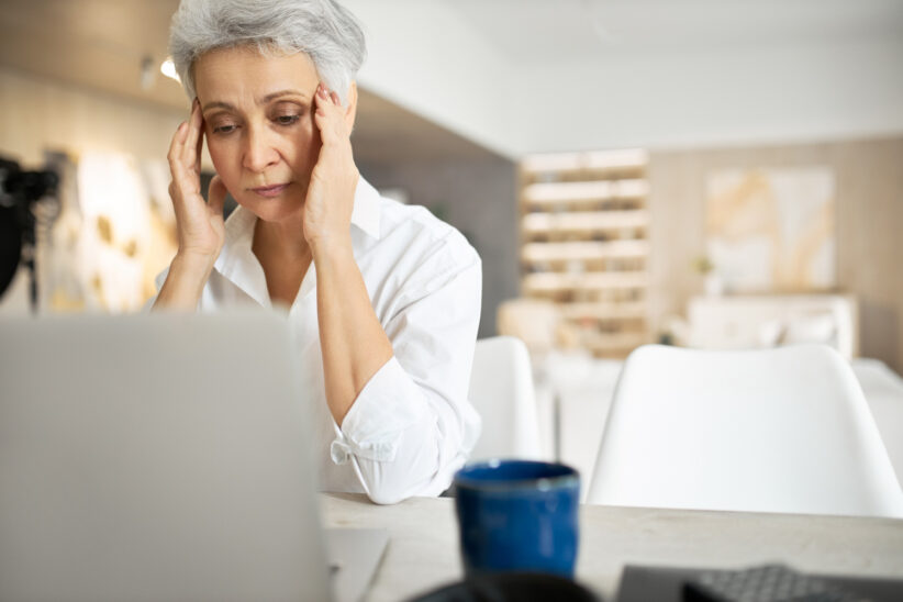 Zmartwiona seniorka siedzi przy laptopie w domu, trzymając dłonie przy skroniach.